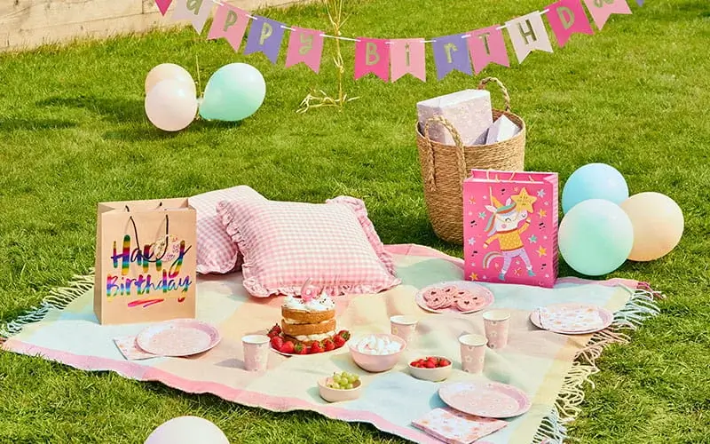 Pastel birthday picnic setup with a birthday cake, balloons, gift bags, pink tableware, cushions, and a Happy Birthday banner on a garden lawn.